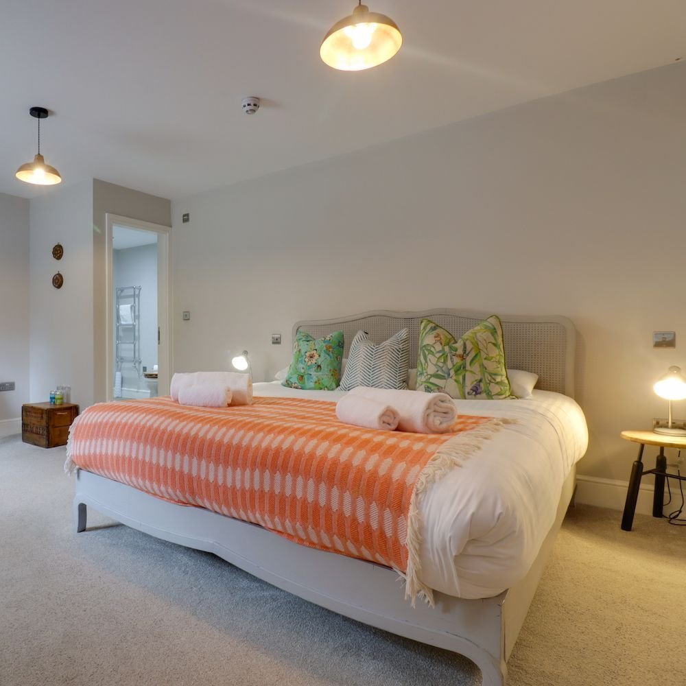 A bright bedroom with a cane headboard, botanical cushions and a coral throw across a white bed, with pendant lights and an ensuite beyond