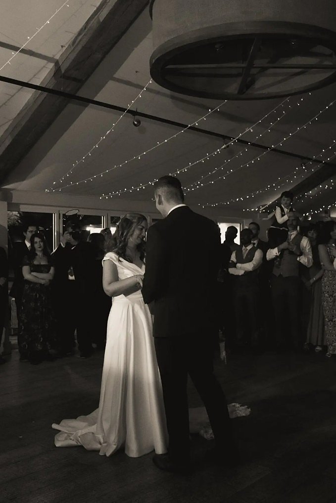 A bride and groom during their first dance in the Pavilion, photographed in black and white, guests standing in a ring around them beneath canopies of fairy lights and the cartwheel chandelier