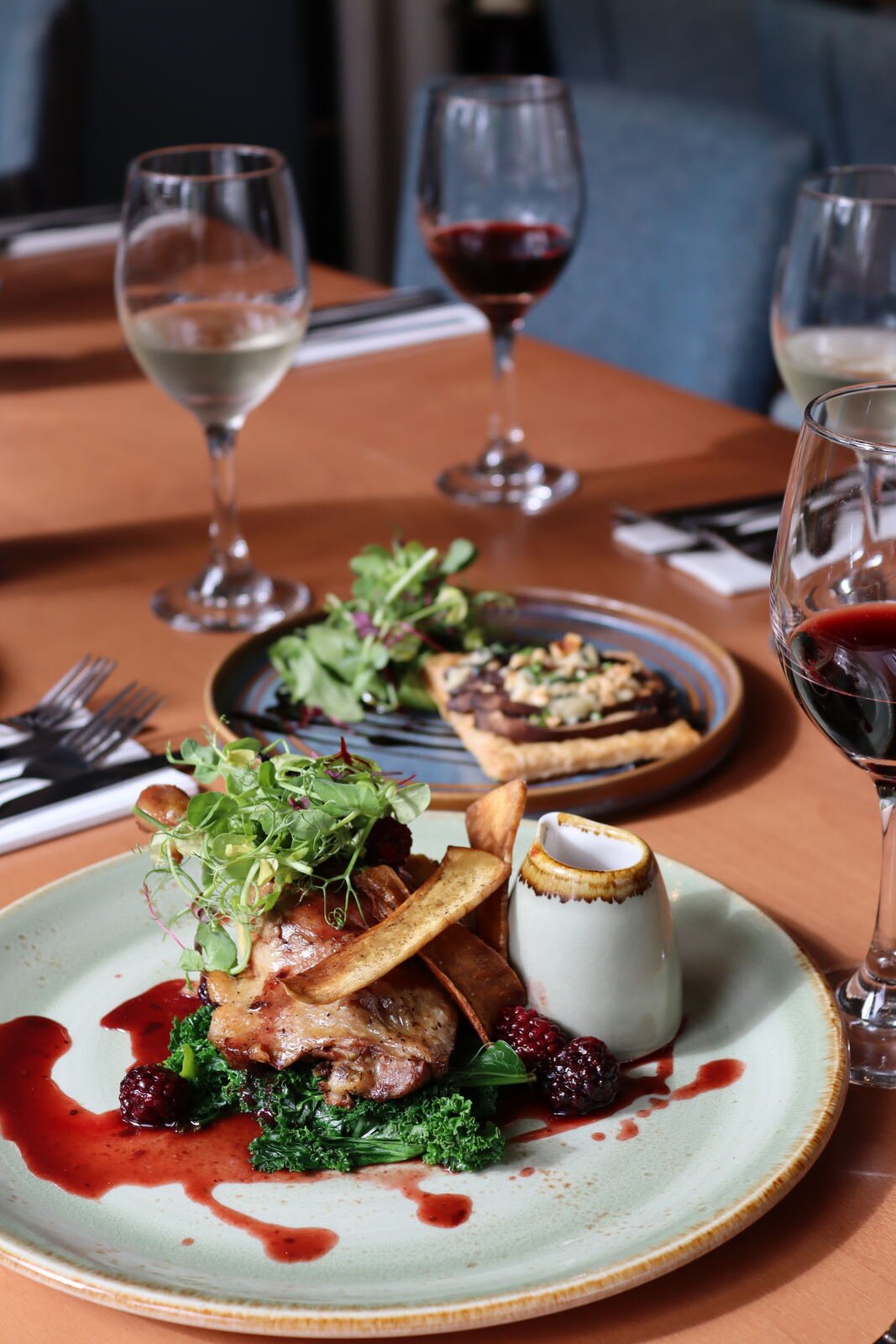Several dishes shared across a restaurant table, with wine glasses, cutlery and upholstered chairs in the background