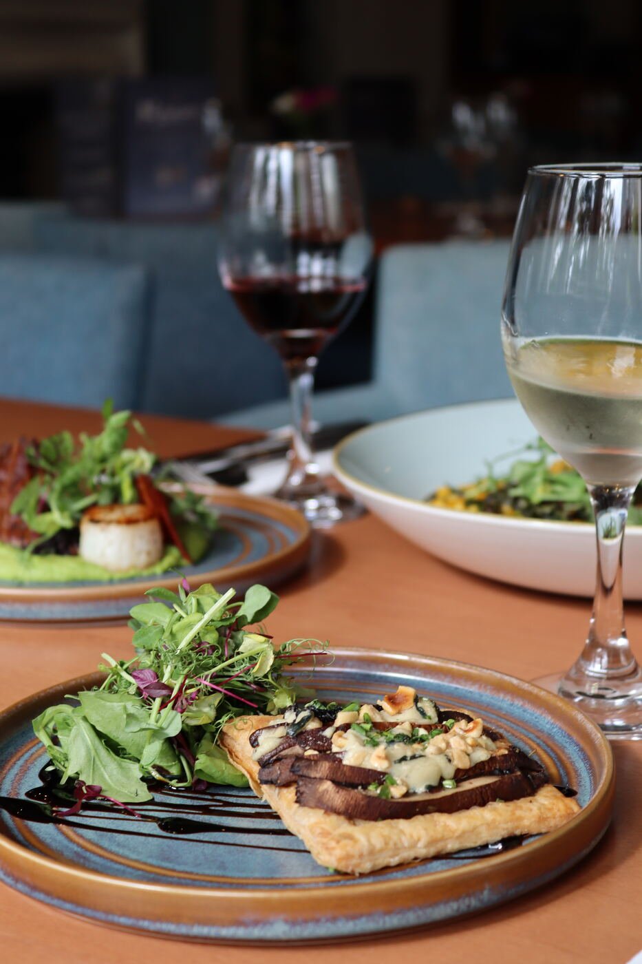 A savoury tart topped with dressed leaves on a rustic glazed plate, photographed on a warm timber table with glasses of red wine