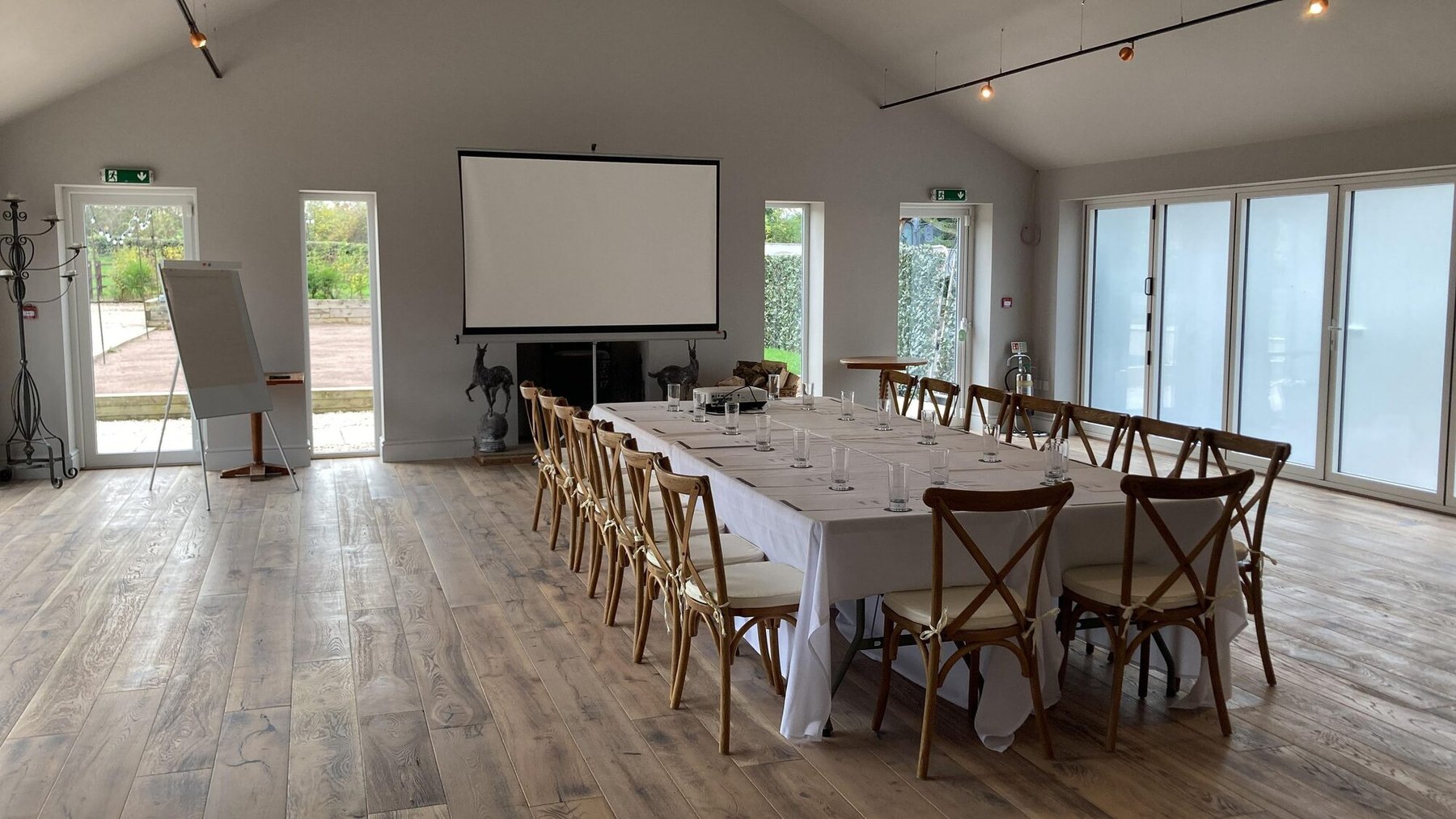The Pavilion laid out for a meeting — a long boardroom table in white linen with glasses and pads set, cross-back chairs down both sides, a projector screen at the far end and a flip chart beside the garden doors