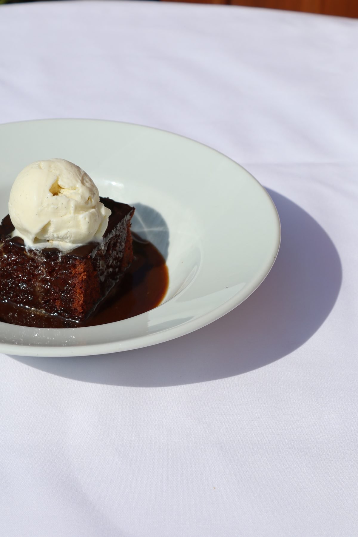 A square of sticky toffee pudding in a shallow white bowl, glossy with toffee sauce and topped with a scoop of vanilla ice cream, on white linen in bright sunlight