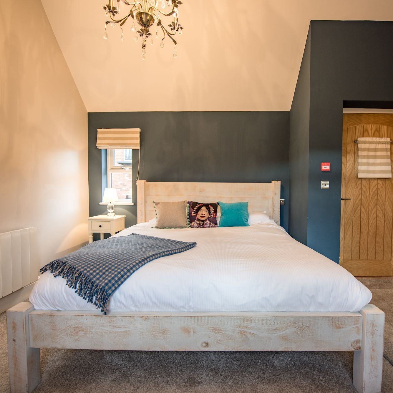 A bedroom with a charcoal feature wall, a whitewashed timber bed, teal and patterned cushions and a chandelier under a sloping ceiling