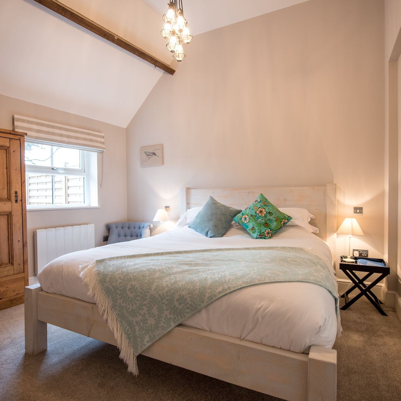 A bedroom under a beamed vaulted ceiling with a caged-bulb pendant light, a whitewashed timber bed frame, sage green throw and cushions, a striped blind at the window and a pine wardrobe beside it