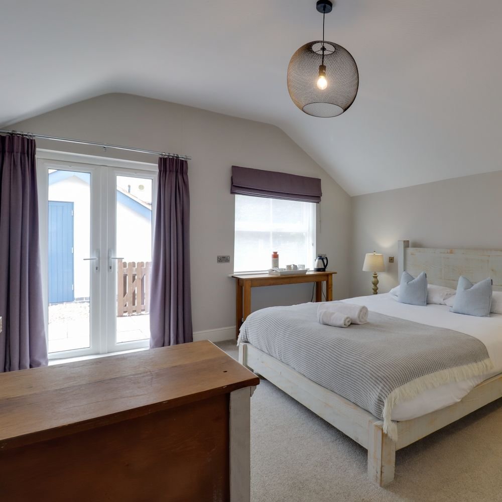 A ground floor bedroom with French doors opening onto a private patio, plum curtains, a whitewashed bed and a wooden blanket box at its foot