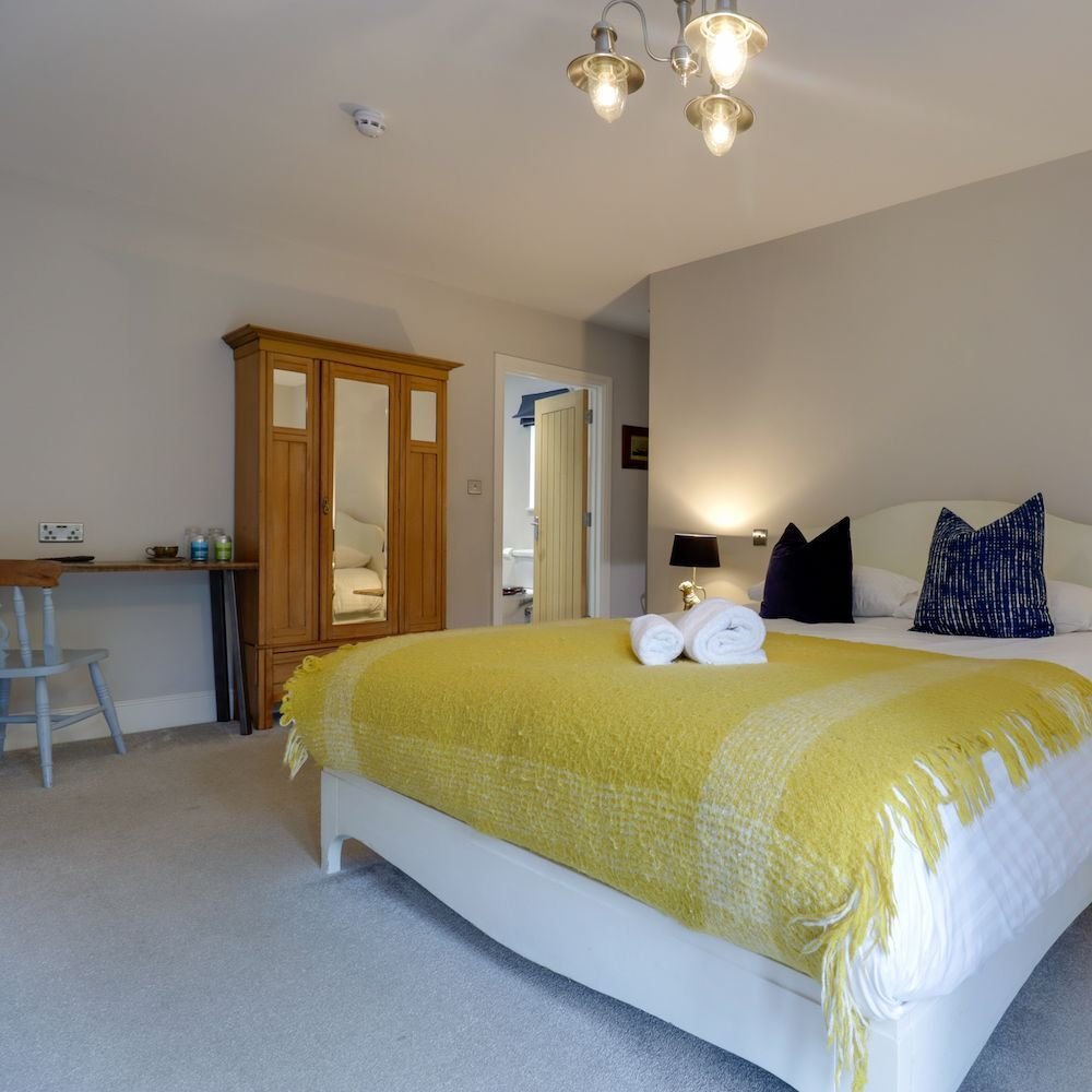 A bedroom with a mustard wool throw and rolled towels across a white bed, navy cushions, a mirrored wardrobe and a small desk under the window