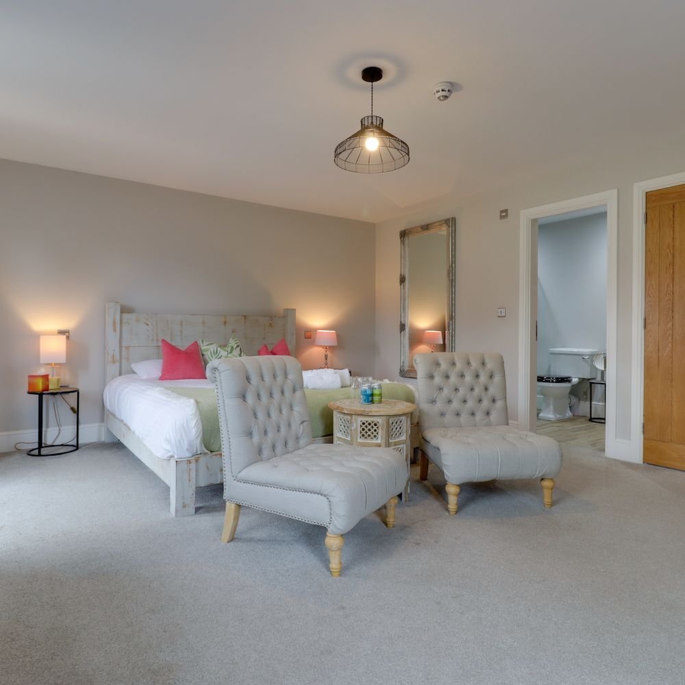 A spacious bedroom with two buttoned armchairs and a small table at the foot of the bed, pink cushions, a tall mirror and an ensuite through the doorway
