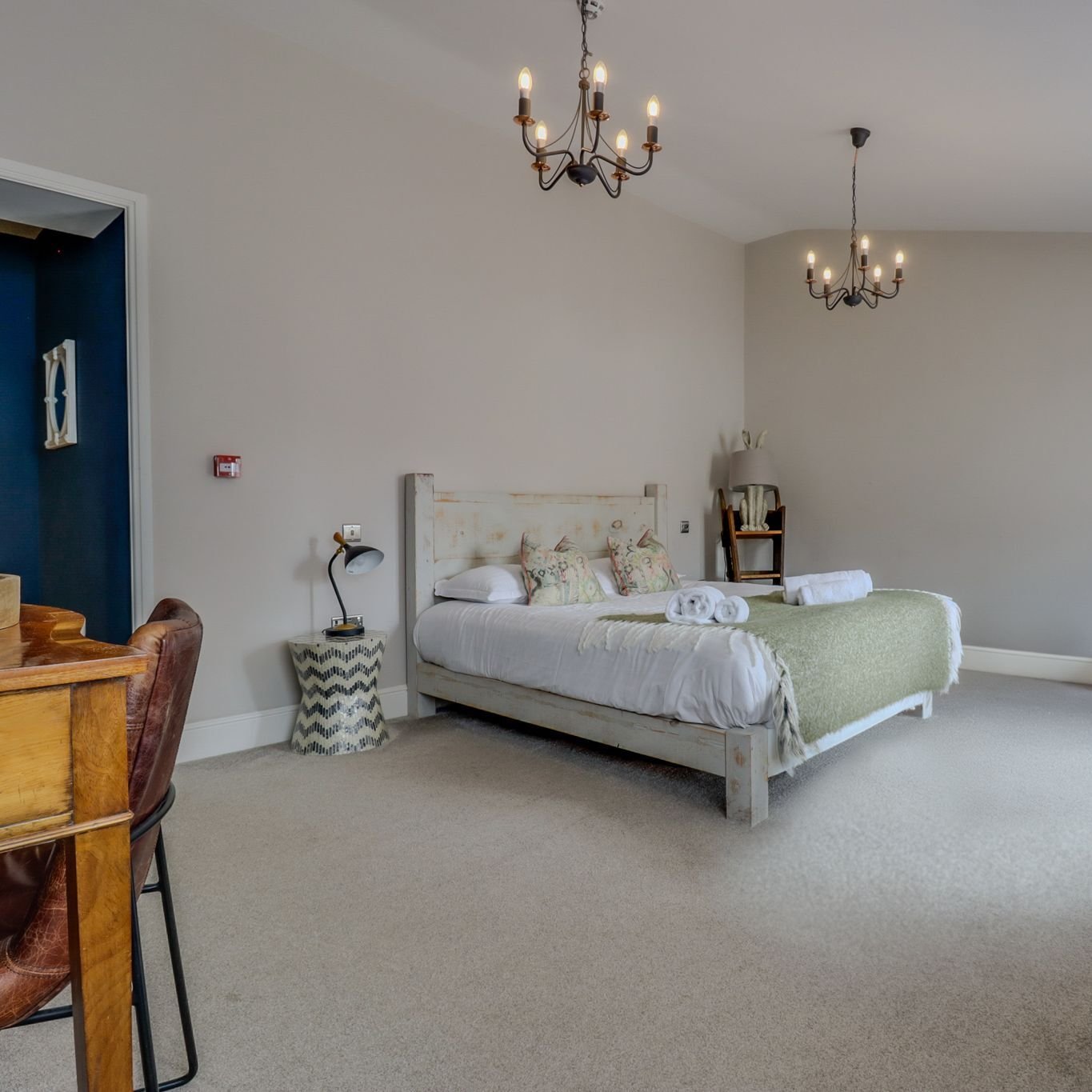 A large bedroom lit by two wrought-iron chandeliers, with a whitewashed bed, a green throw, a leather chair at a wooden desk and a deep blue alcove beyond