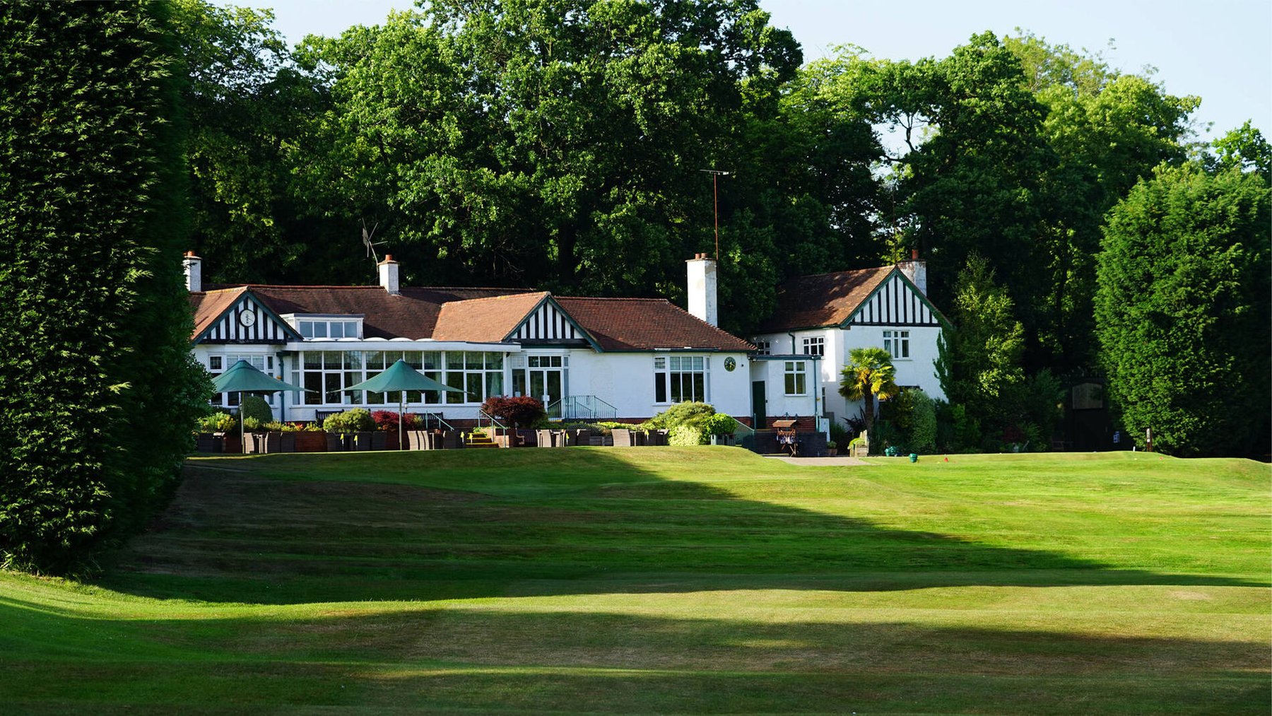 A golf clubhouse in black and white timber seen across a wide green fairway, framed by mature trees