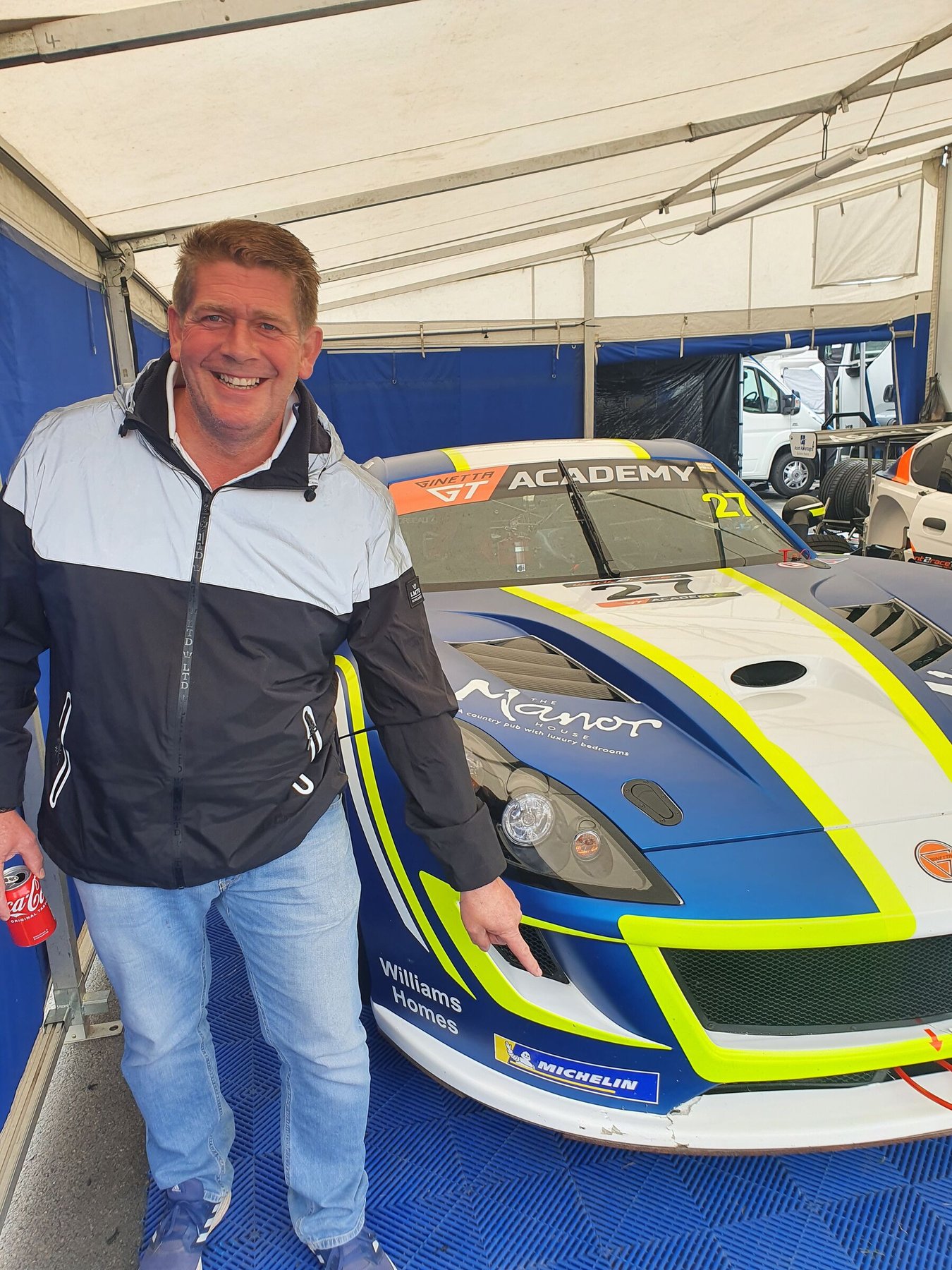 Steve standing beside his blue and yellow GT racing car in the paddock awning, the Manor House name on the bodywork