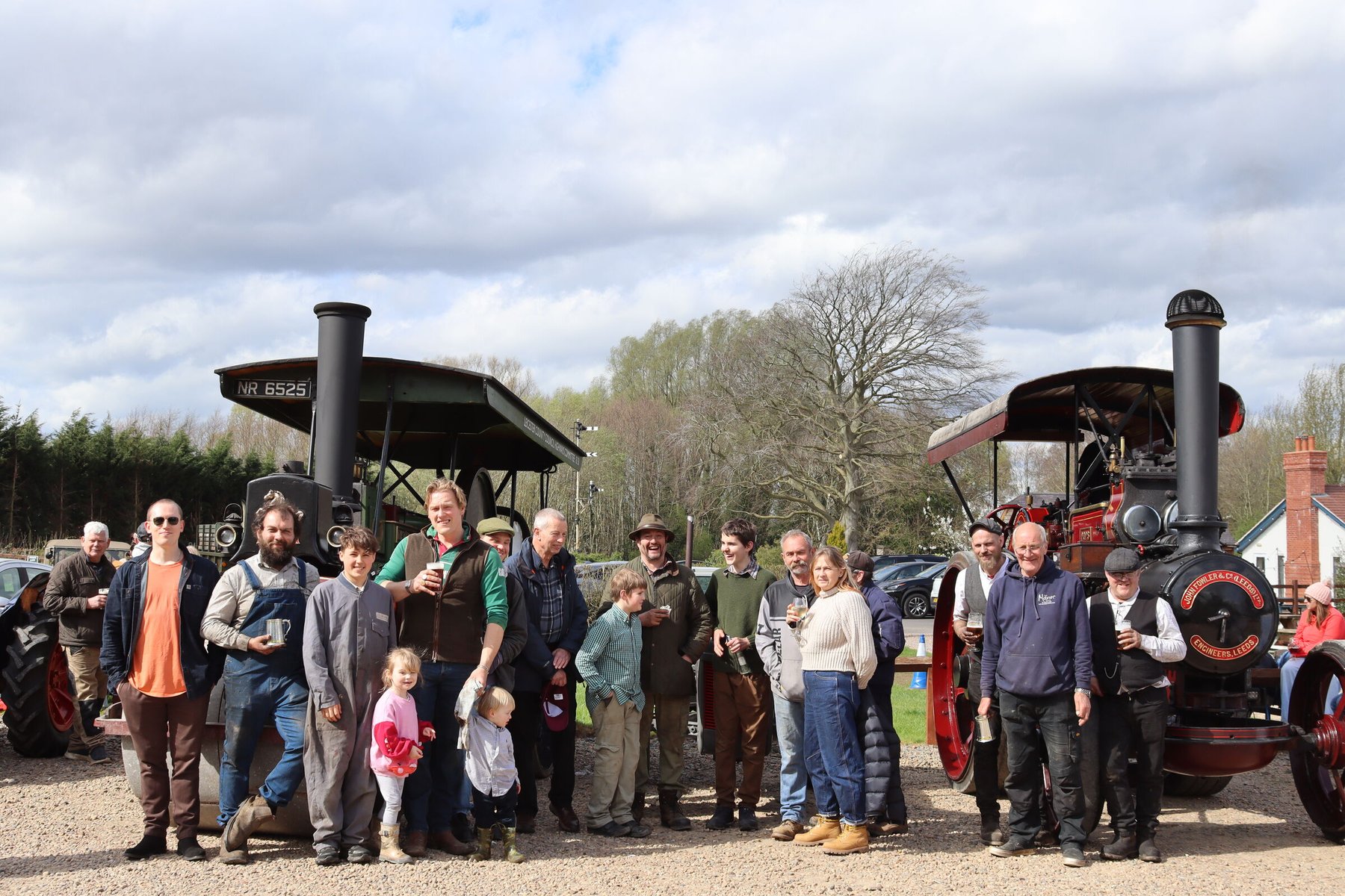 A group of locals of all ages gathered outside with pints in hand between two restored steam traction engines on a bright spring day