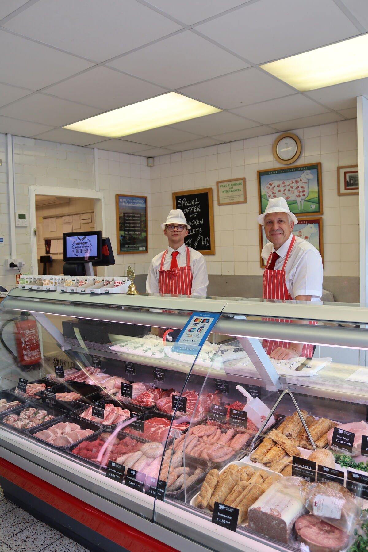 Two butchers in white hats and red striped aprons behind a full counter of meat, sausages and pies in their shop