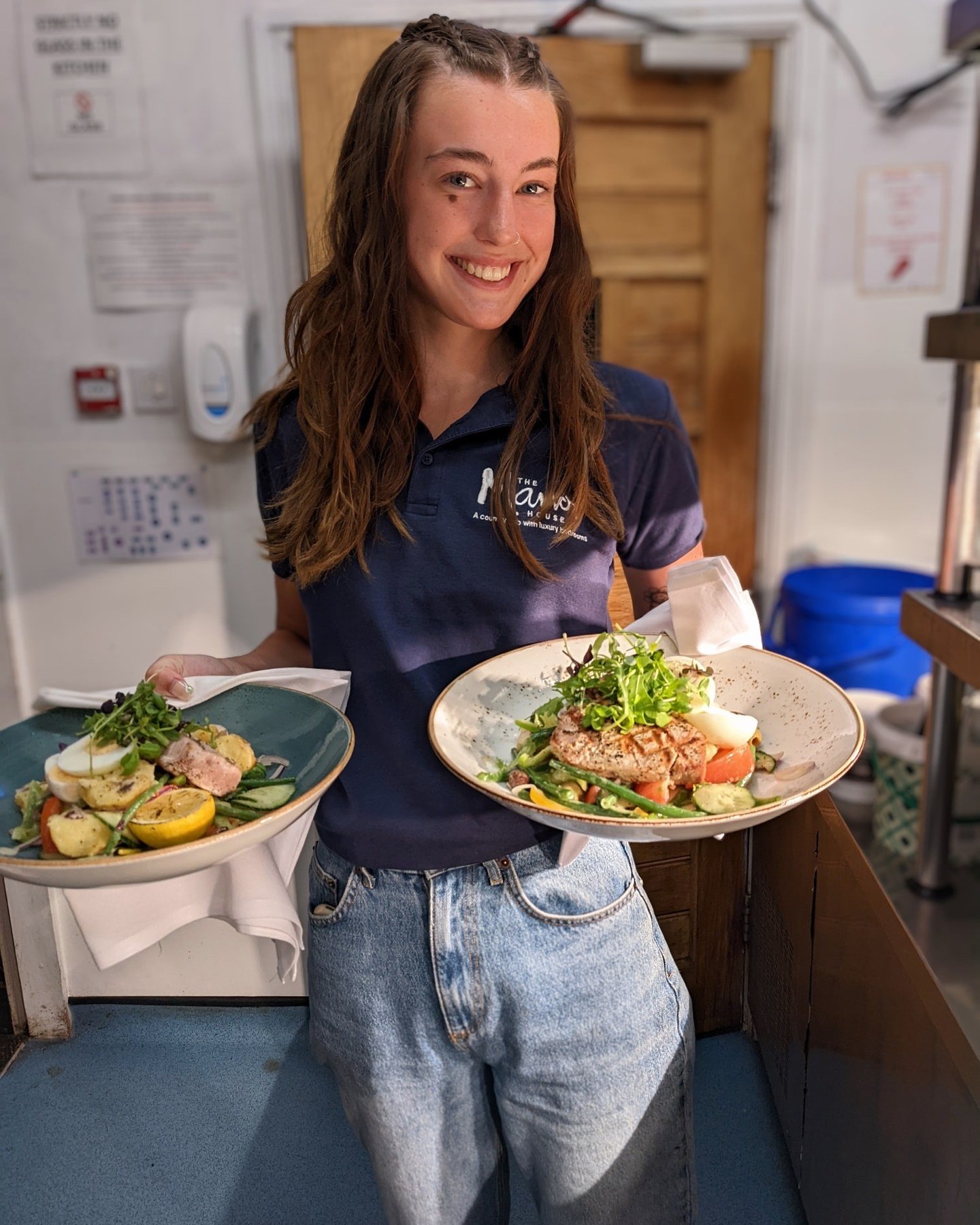 A member of the front of house team in a Manor House polo shirt carrying two plated dishes out of the kitchen, smiling at the camera