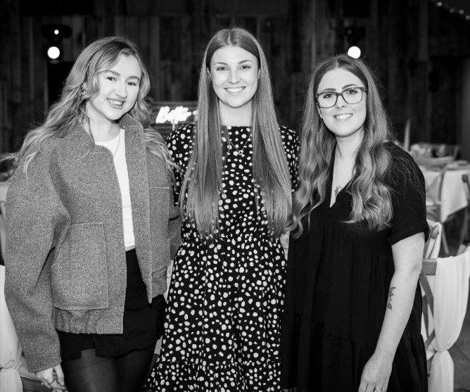 Three members of the team standing together in the Pavilion, photographed in black and white, laid tables behind them