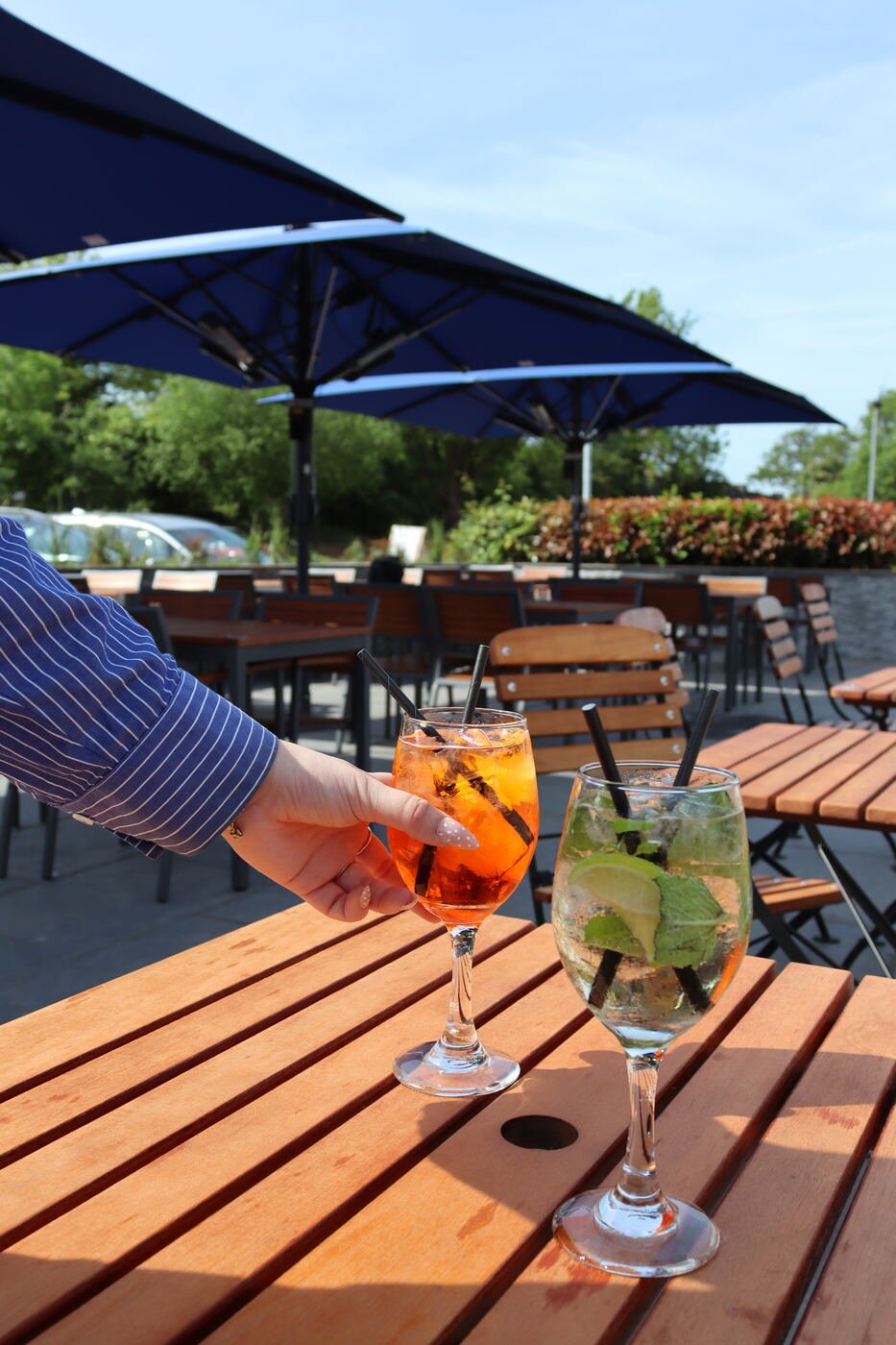 A member of staff placing two cocktails onto a wooden terrace table beneath a navy parasol