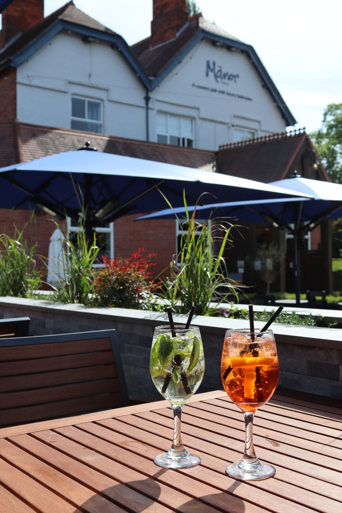 Two cocktails on a wooden terrace table under navy parasols, with the cream and timber frontage of the building behind on a clear sunny day