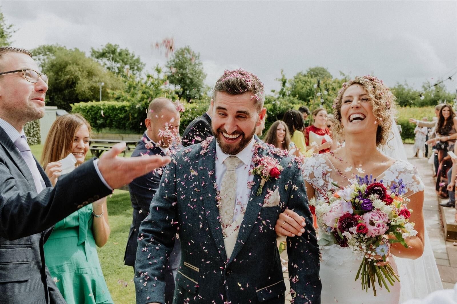 A newly married couple walking out through a shower of pink petal confetti, both laughing, guests lining the path on either side and the bride holding a bright mixed bouquet
