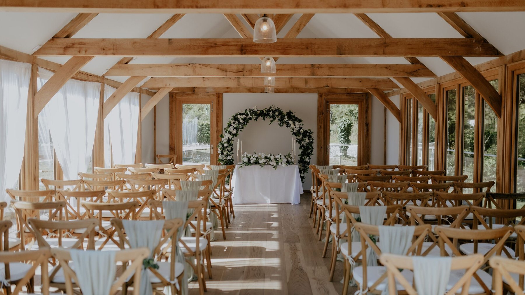 The Glade House set for a ceremony — an oak-framed room with exposed trusses and glazing down both sides, rows of wooden cross-back chairs dressed in sage chiffon, and a circular arch of white flowers and eucalyptus behind the registrar's table