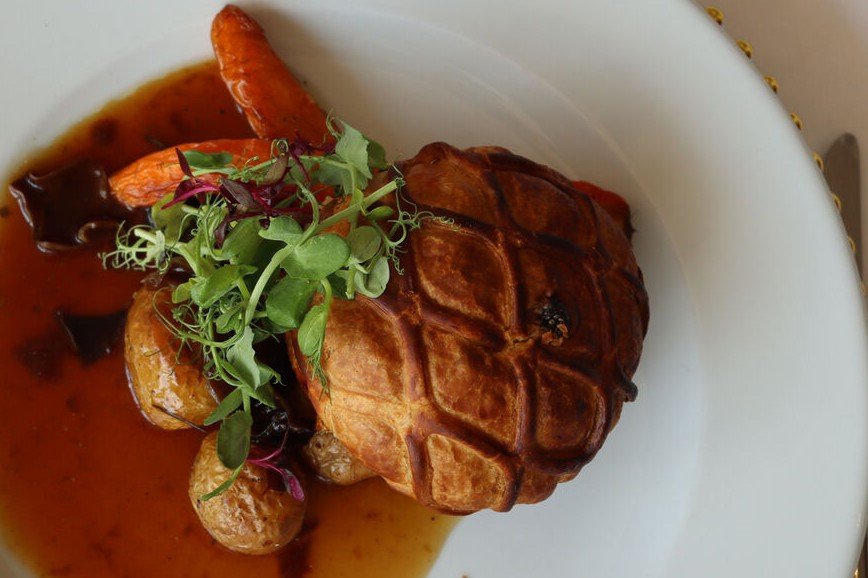 A beef wellington in lattice-scored golden puff pastry, with roasted carrots, new potatoes, a dark red wine sauce and a tangle of pea shoots, on white china with a gold-beaded charger