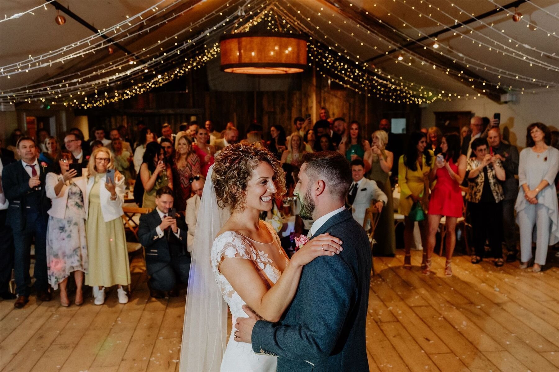 A couple's first dance on a wooden floor beneath canopies of fairy lights, with guests gathered around watching