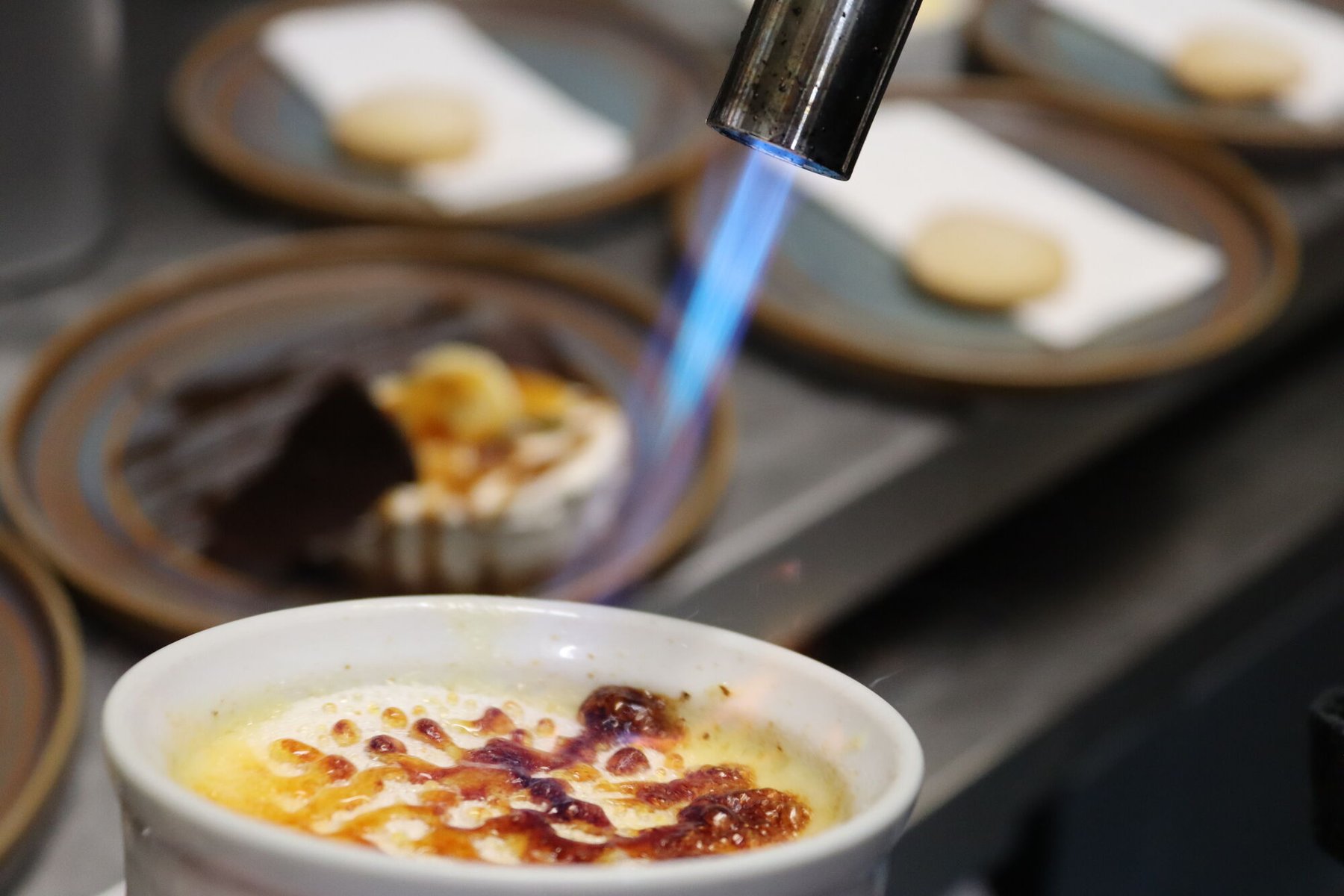 A crème brûlée being caramelised with a blowtorch on the pass, further desserts plated and waiting behind